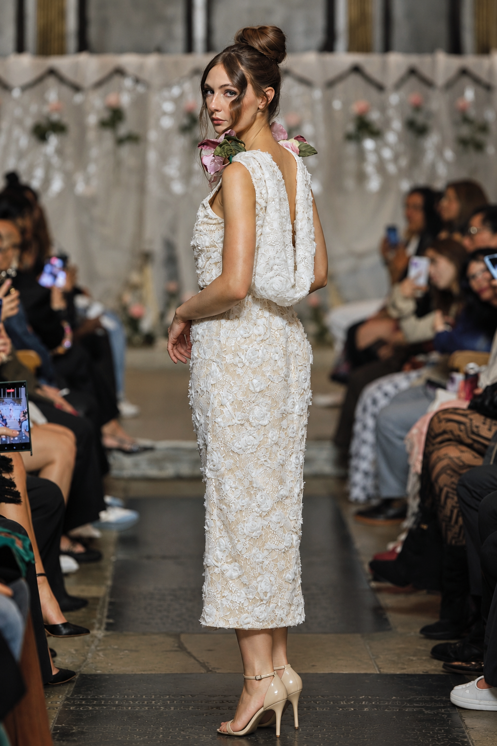 A model on a runway wearing a sleeveless, textured cream lace midi dress with a draped shoulder detail and nude high heels, with seated spectators on both sides.