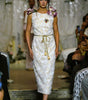 A model walks the runway wearing a sleeveless white lace midi dress with a gold tie belt, floral shoulder details, and a brooch at the neckline.