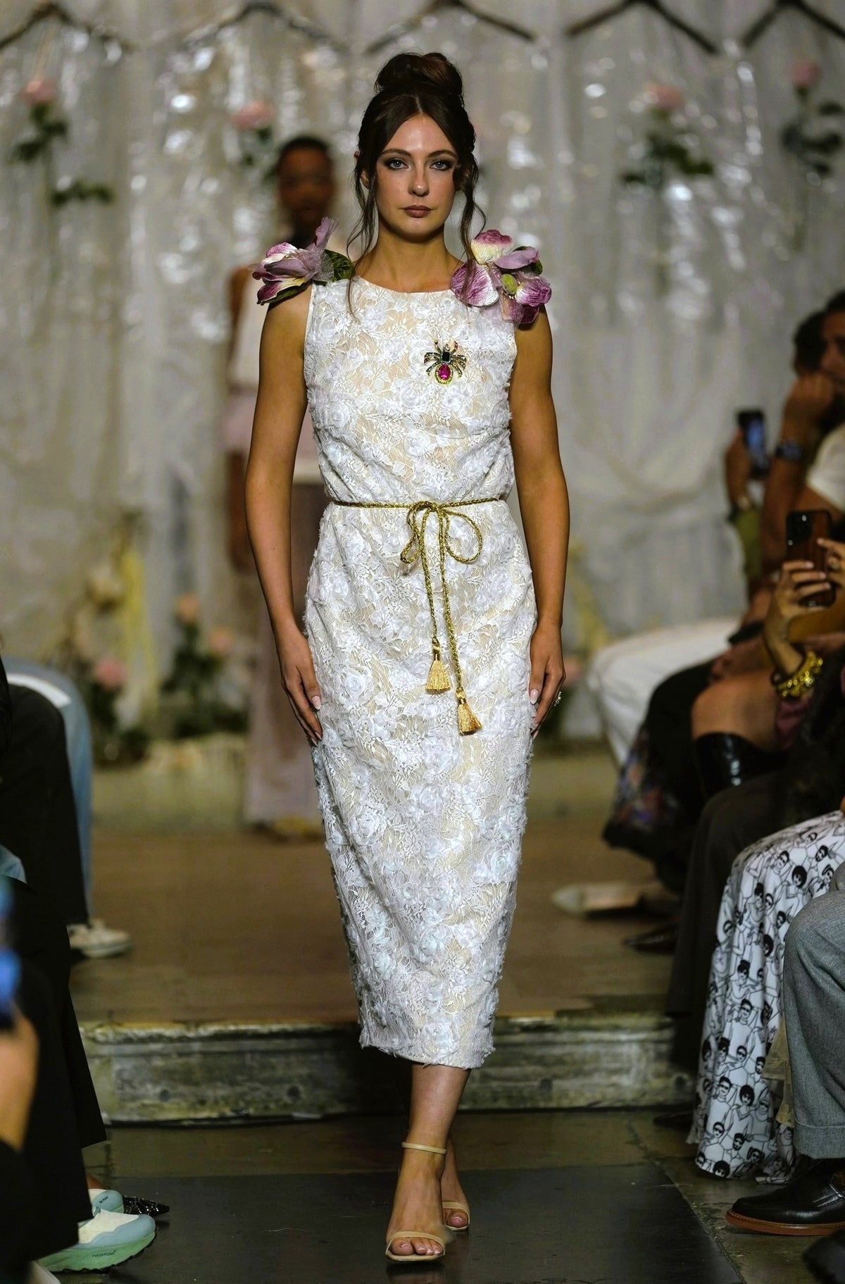 A model walks the runway wearing a sleeveless white lace midi dress with a gold tie belt, floral shoulder details, and a brooch at the neckline.