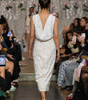 A model walks down a fashion runway wearing a sleeveless, deep V-back lace dress with floral applique and small pink flower accents on the shoulders. The runway is lined with seated spectators holding up phones to capture the show.