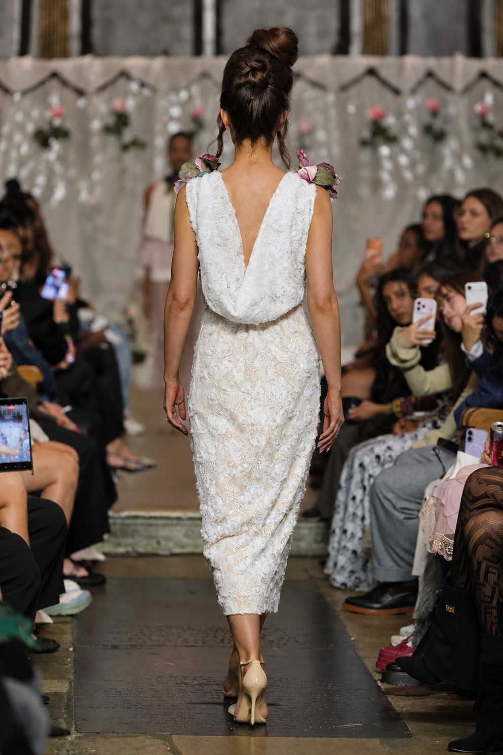 A model walks down a fashion runway wearing a sleeveless, deep V-back lace dress with floral applique and small pink flower accents on the shoulders. The runway is lined with seated spectators holding up phones to capture the show.