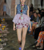 Model walking on a runway wearing a floral dress and accessories.