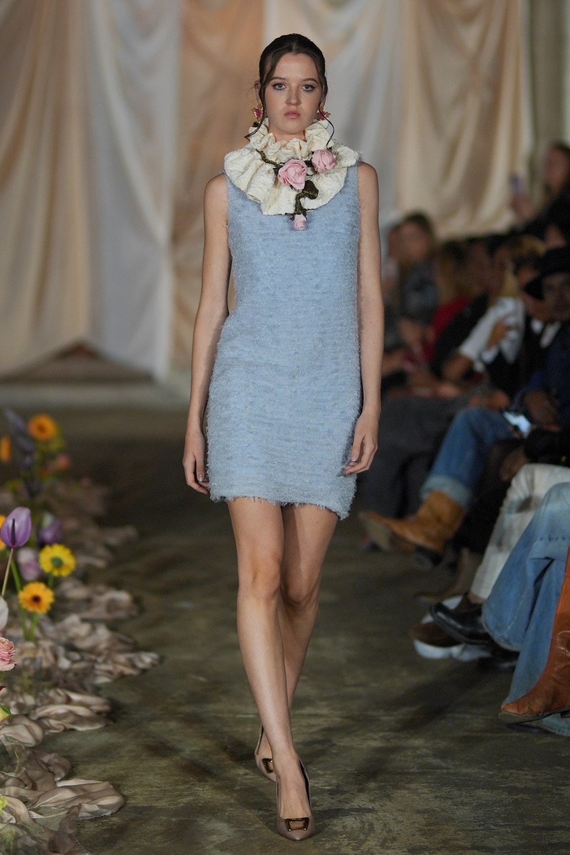 Model wearing a light blue dress with floral accessories on a runway.