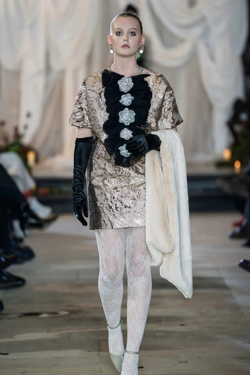 Model on a runway wearing a detailed outfit with floral elements and gloves.