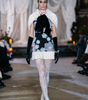 Model walking on a runway wearing a floral dress with white fur accents and black gloves.