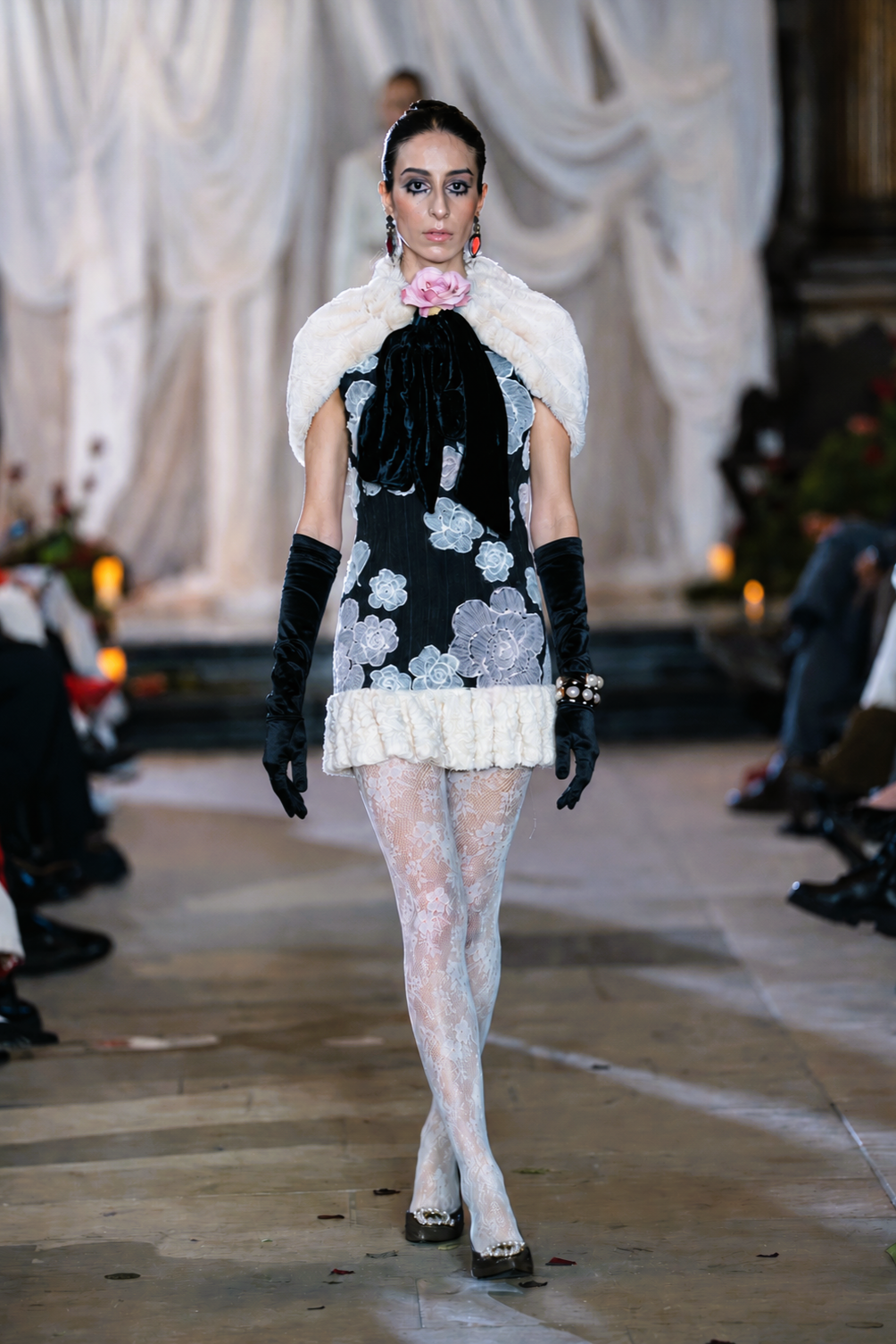 Model walking on a runway wearing a floral dress with white fur accents and black gloves.