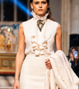 Model walking on a runway wearing a white outfit with a patterned scarf.
