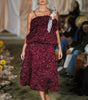 Model wearing a burgundy floral dress on a runway with an audience.