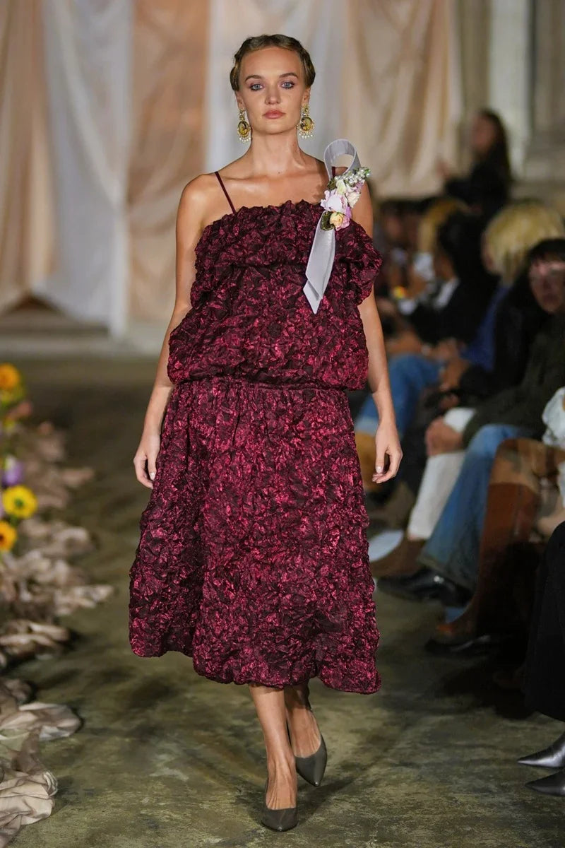 Model wearing a burgundy floral dress on a runway with an audience.