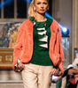 Model walking on a fashion show runway wearing a green top, orange coat, beige pants, and brown boots.
