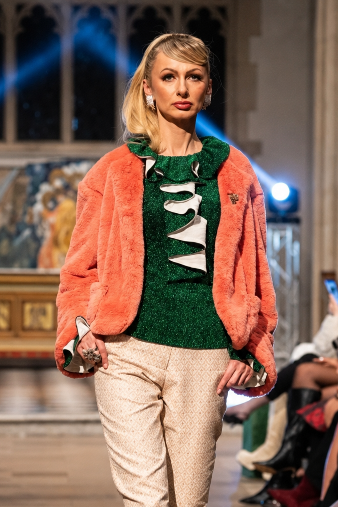 Model walking on a fashion show runway wearing a green top, orange coat, beige pants, and brown boots.