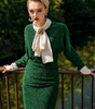 Woman in a green dress with a white scarf standing outdoors with greenery in the background