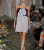Model walking on a runway wearing a burgundy top and beige skirt with a white garment over the top.