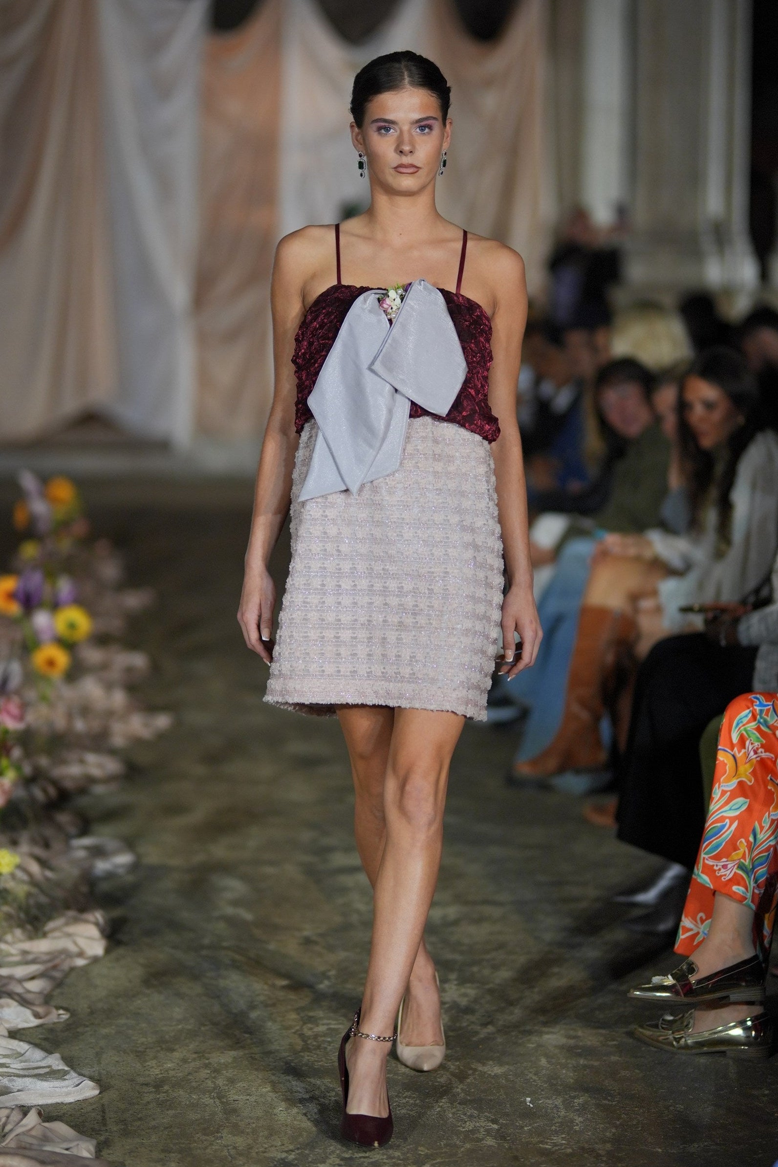 Model walking on a runway wearing a burgundy top and beige skirt with a white garment over the top.