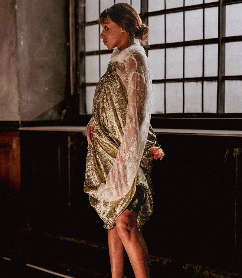 Woman in a gold and white dress standing in a dimly lit room with large windows.