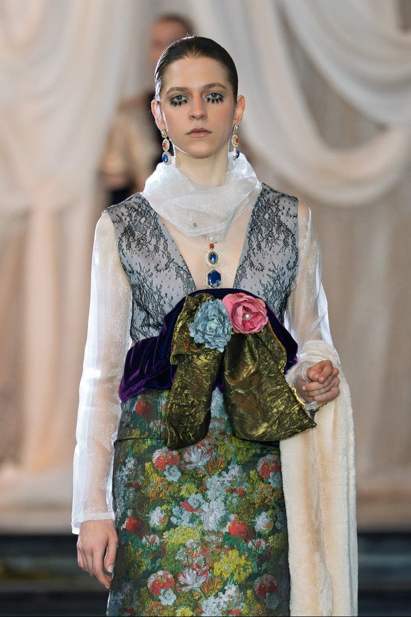 Model walking on a fashion runway wearing a floral dress and patterned vest.