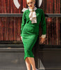 Woman in a green outfit with a white scarf standing against a textured wall.