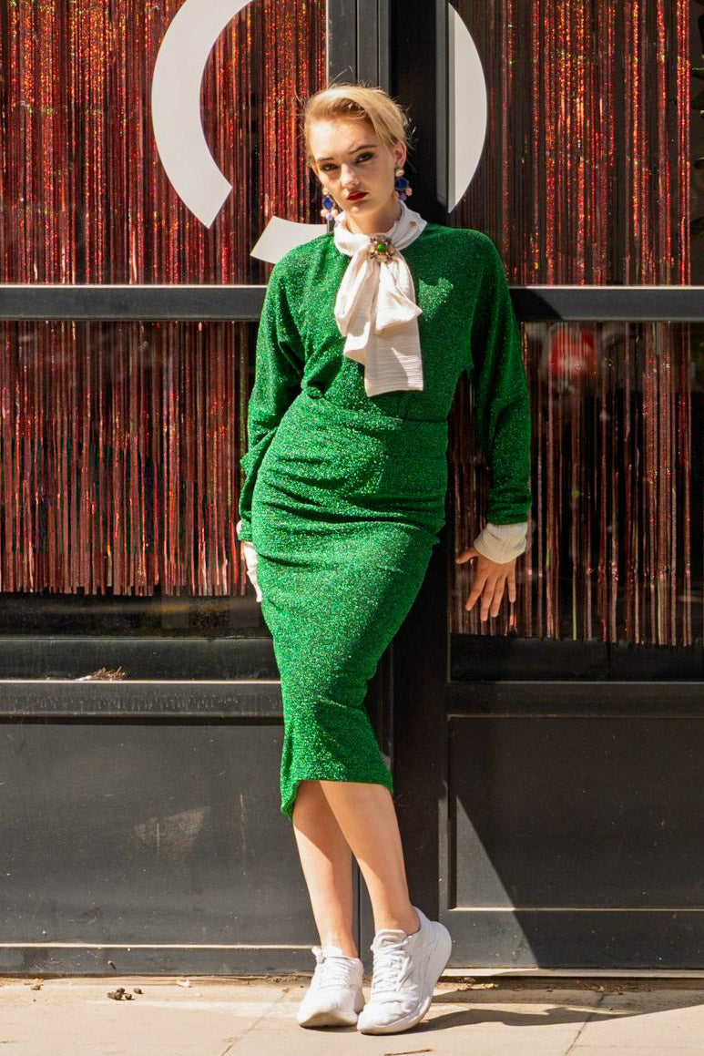 Woman in a green outfit with a white scarf standing against a textured wall.