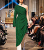 Model wearing a green one-shoulder dress on a fashion runway with an audience.