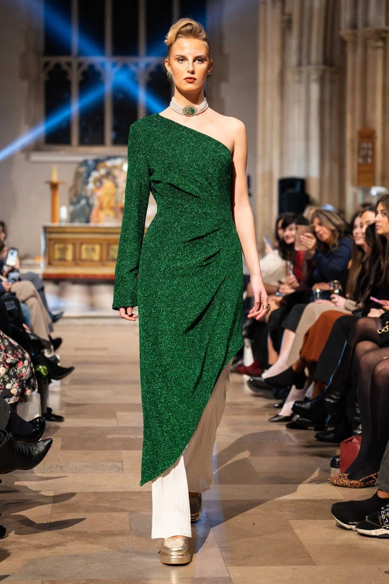 Model wearing a green one-shoulder dress on a fashion runway with an audience.