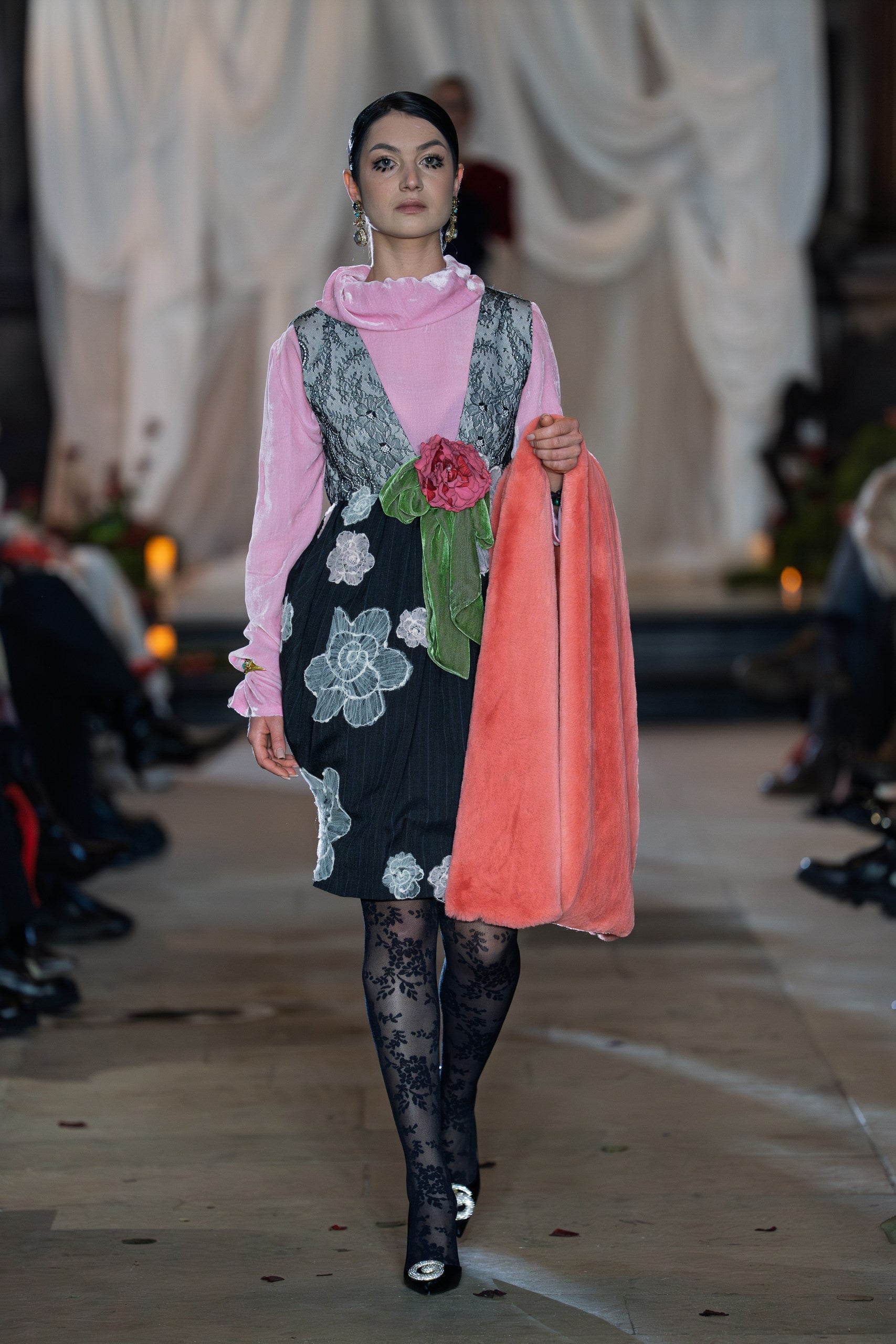 Model walking on a runway wearing a floral dress with a pink shawl.