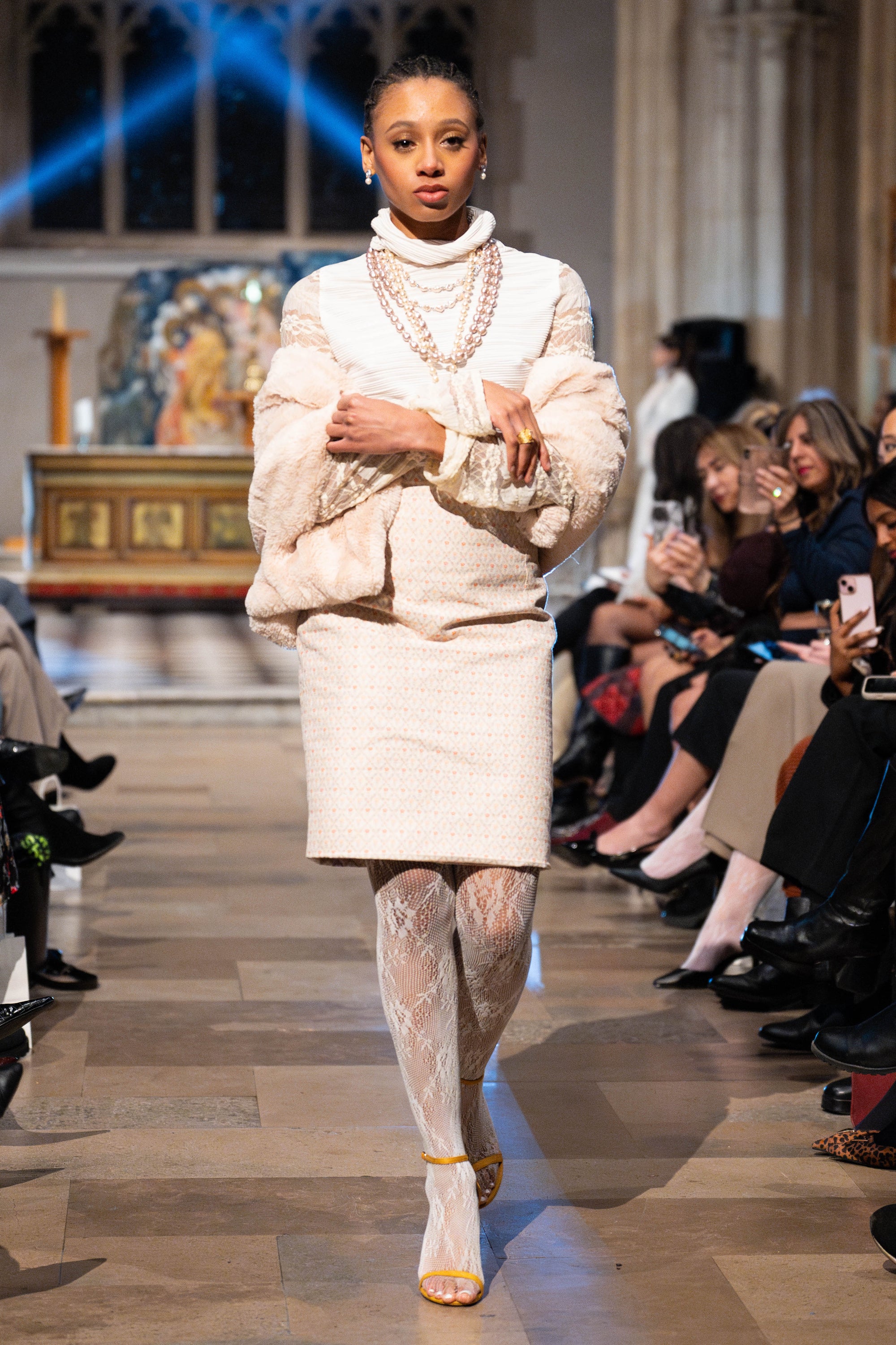 Model walking on a fashion show runway wearing a white outfit with lace details.