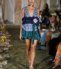Model walking on a runway wearing a floral dress with a green belt.