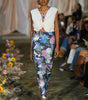 Model walking on a runway wearing a white top and colorful floral pants.