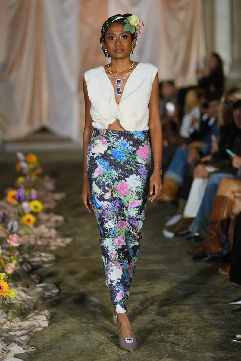 Model walking on a runway wearing a white top and colorful floral pants.