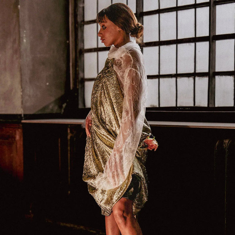 Woman in a gold and white dress standing in a dimly lit room with large windows.