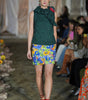 Model walking on a fashion show runway wearing a green top and colorful floral skirt.