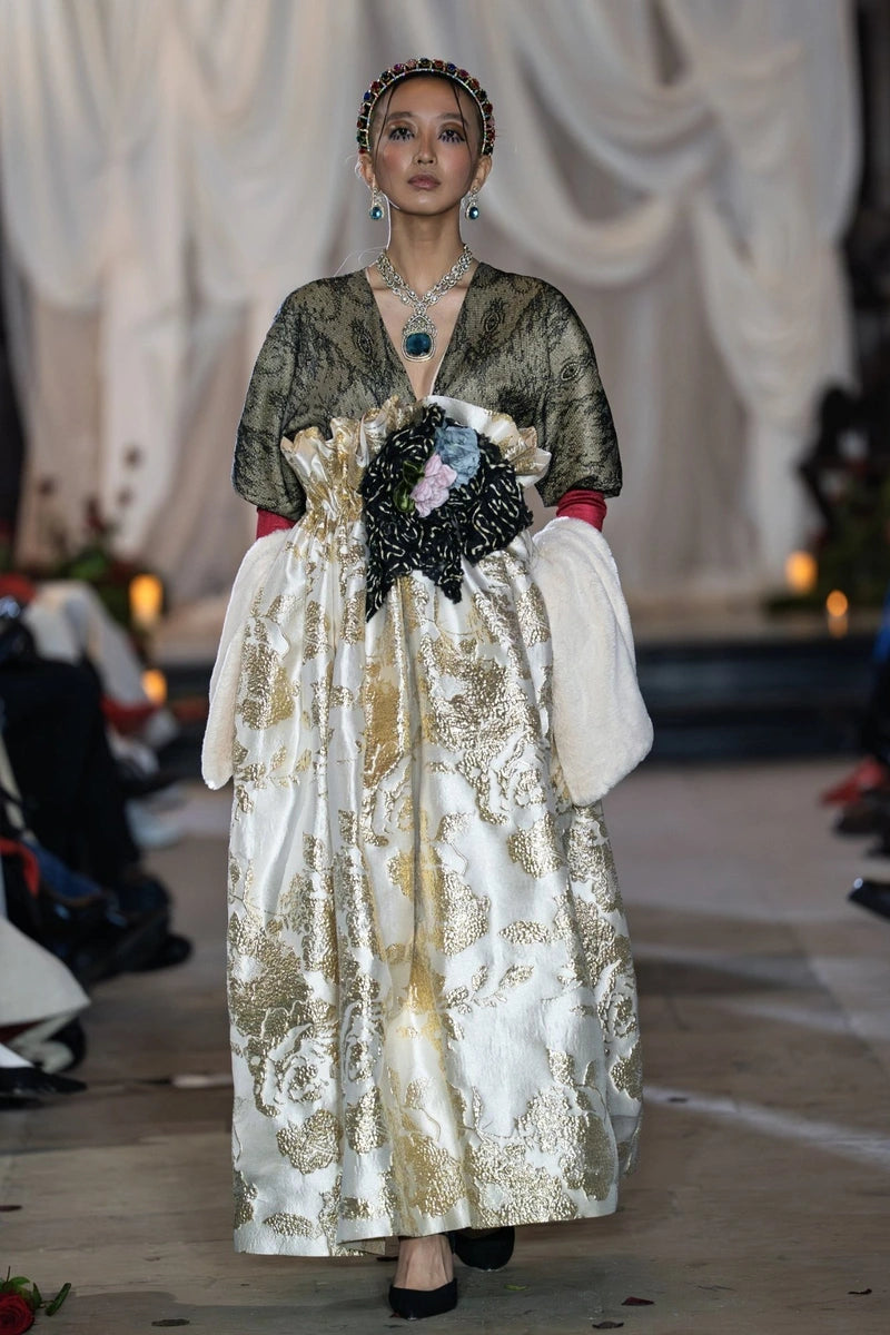 Model wearing a long, elegant dress with gold and white patterns on a runway.