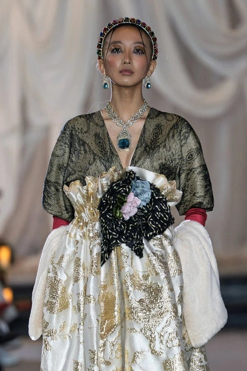 Model wearing a long, elegant dress with gold and white patterns on a runway.