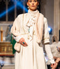 Model wearing a white outfit on a runway with an audience in a formal setting.