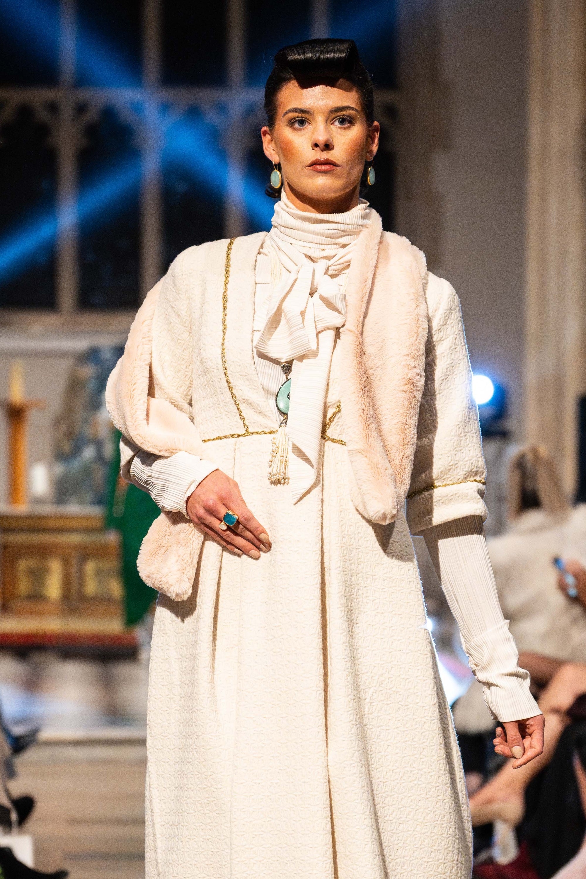 Model wearing a white outfit on a runway with an audience in a formal setting.