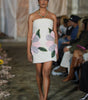 Model walking on a runway wearing a floral dress with an audience watching.