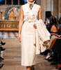 Model walking on a runway wearing a white outfit with a patterned scarf.