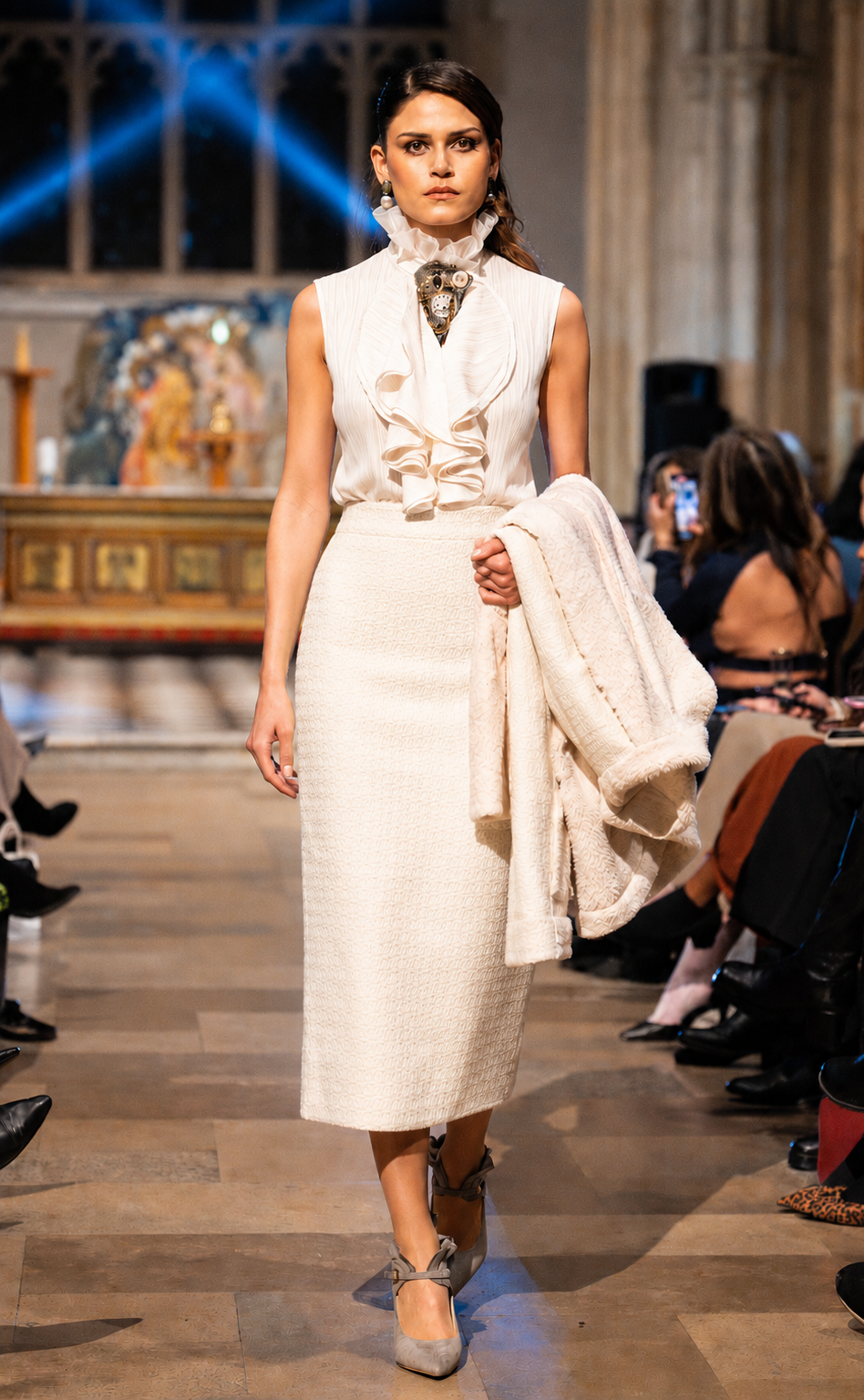 Model walking on a runway wearing a white outfit with a patterned scarf.