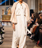 Model walking on a fashion show runway wearing a white outfit with a beige shawl.