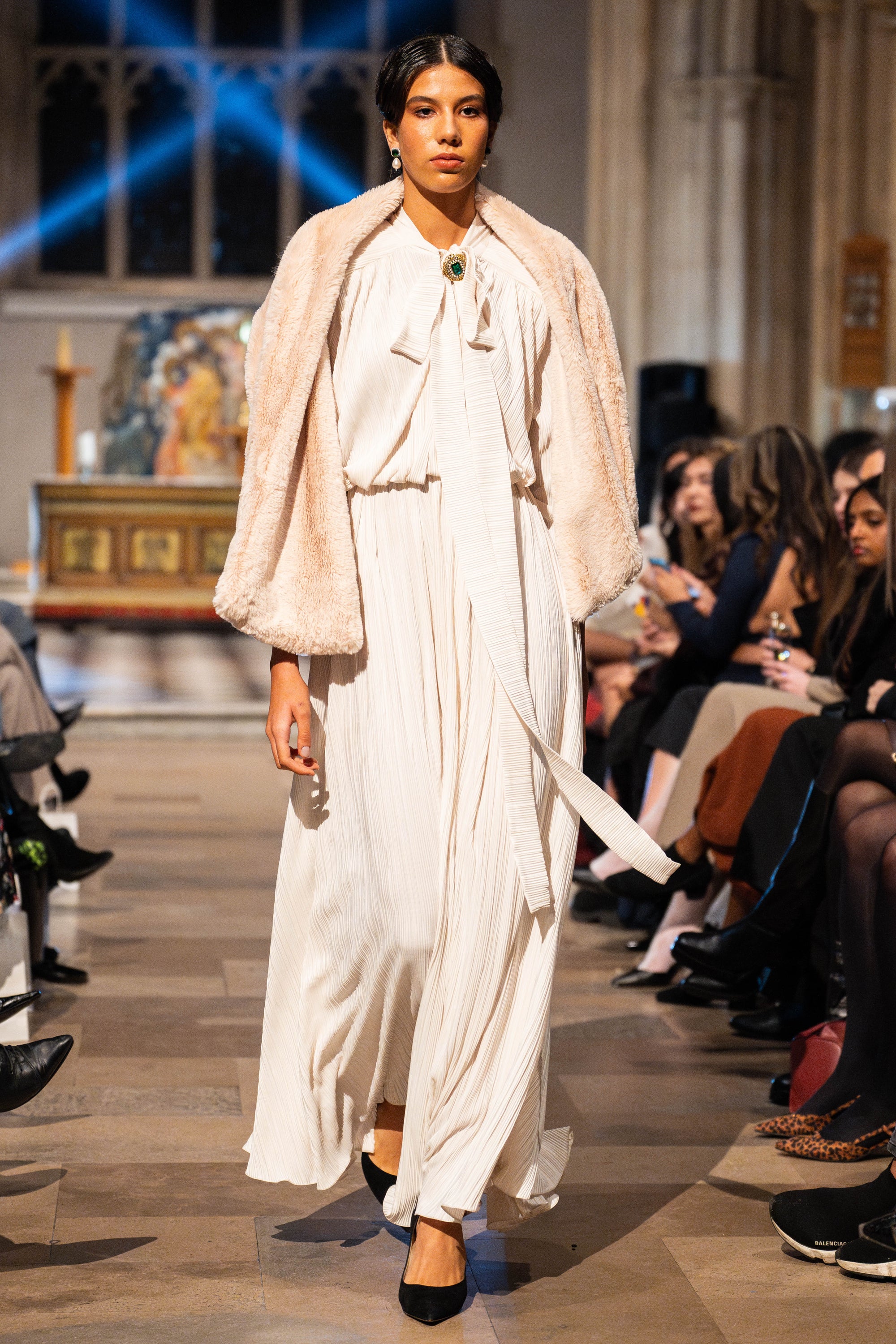 Model walking on a fashion show runway wearing a white outfit with a beige shawl.