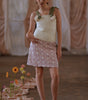 Model wearing a white top and pink skirt with floral patterns against a draped curtain background.