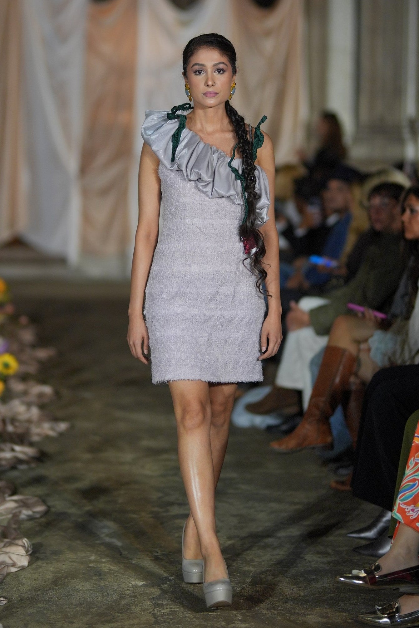 Model walking on a runway wearing a gray dress with ruffled details.
