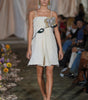 Model walking down a runway in a white dress with floral details, surrounded by an audience.