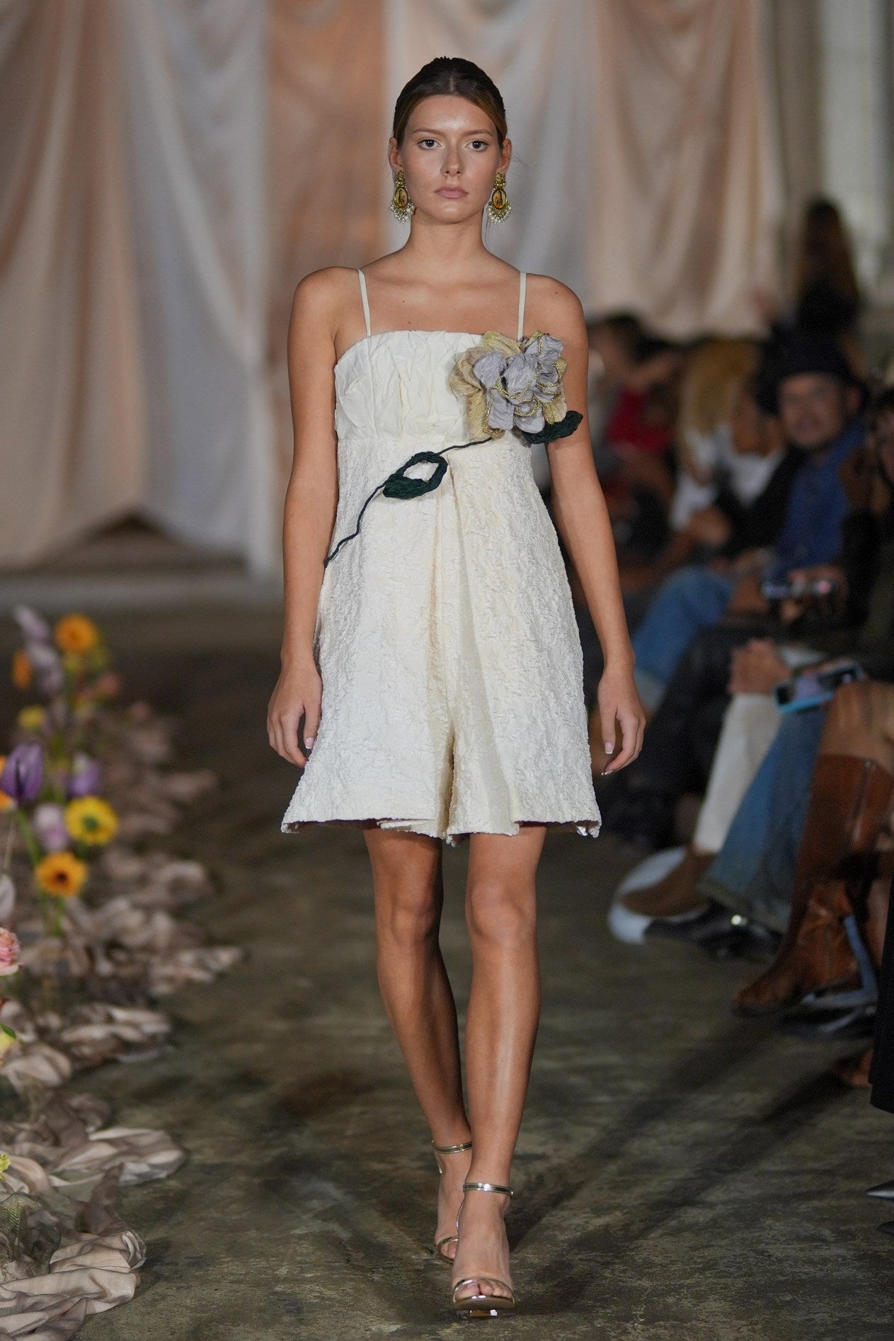 Model walking down a runway in a white dress with floral details, surrounded by an audience.