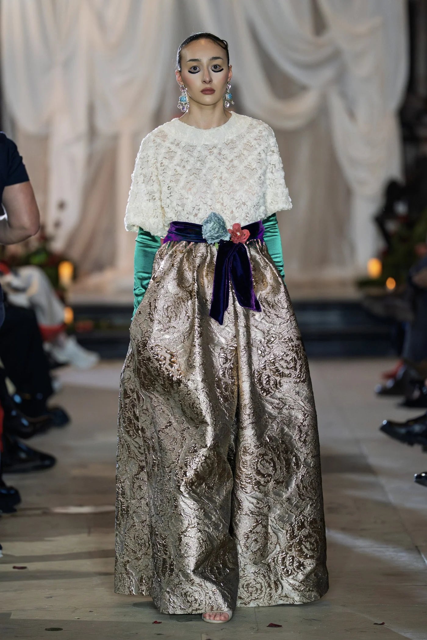 Model wearing a white lace top and patterned pants on a runway with a blurred background