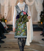 Model walking on a runway wearing a colorful floral dress with a white coat.