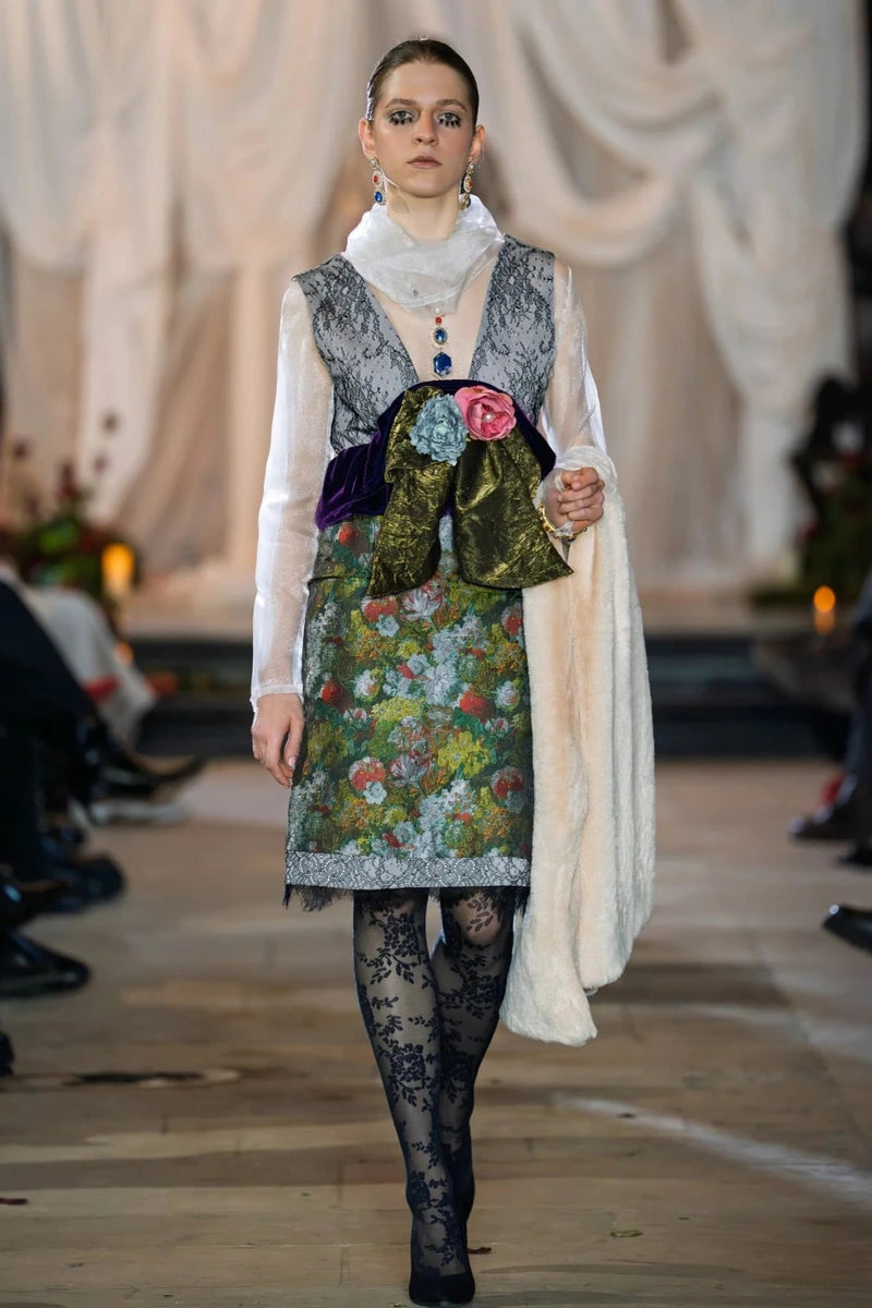 Model walking on a runway wearing a colorful floral dress with a white coat.