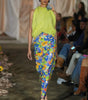 Model wearing a bright green top and colorful pants on a fashion runway.
