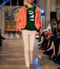 Model walking on a runway wearing a bright orange fur coat, green sweater, and beige pants.
