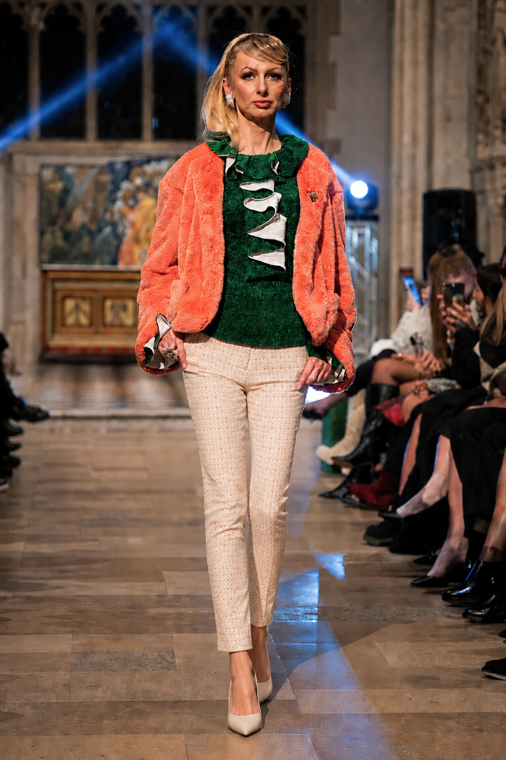 Model walking on a runway wearing a bright orange fur coat, green sweater, and beige pants.